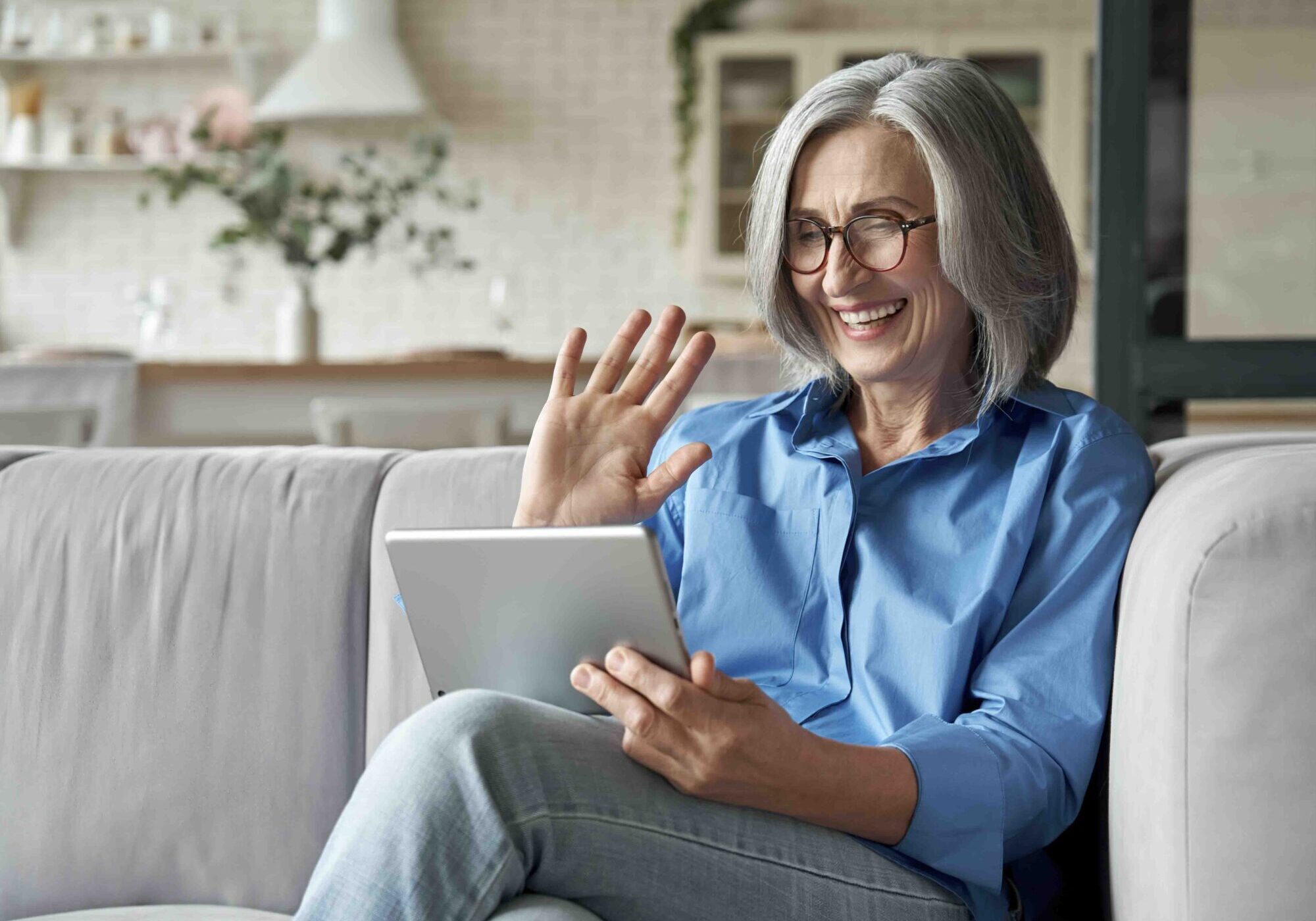 Happy 60s older mature middle aged adult woman waving hand holding digital tablet computer video conference calling by social distance virtual family online chat meeting sitting on couch at home.